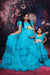 Two women in matching teal dresses with floral accessories against a decorative floral backdrop.